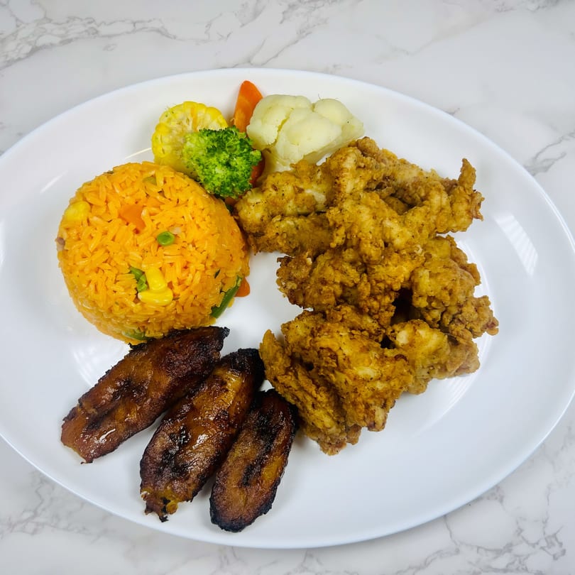 A white plate holds fried chicken, yellow rice, plantains, and steamed vegetables.