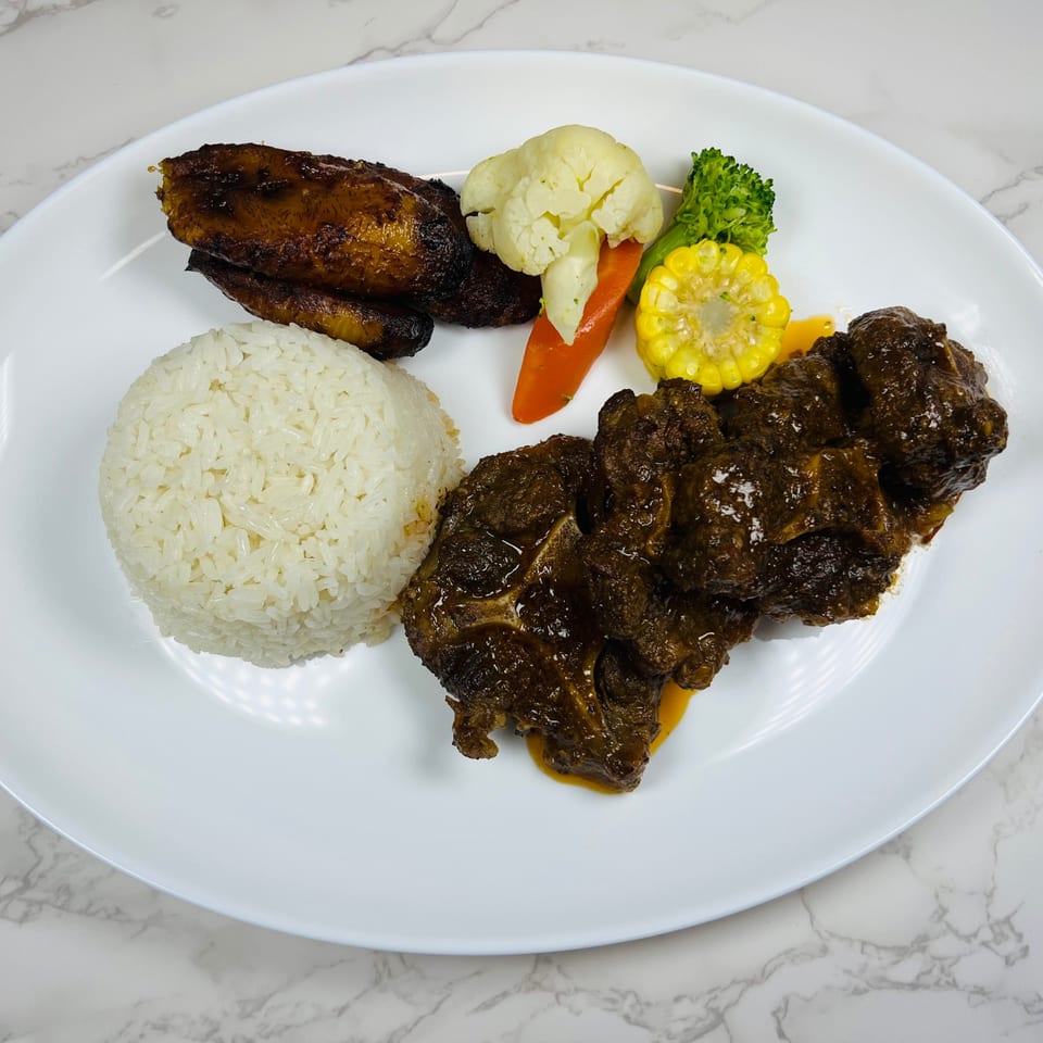 Oxtail stew with white rice, fried plantains, and steamed vegetables on a white plate.