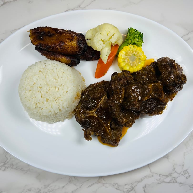 Oxtail stew with white rice, fried plantains, and steamed vegetables on a white plate.